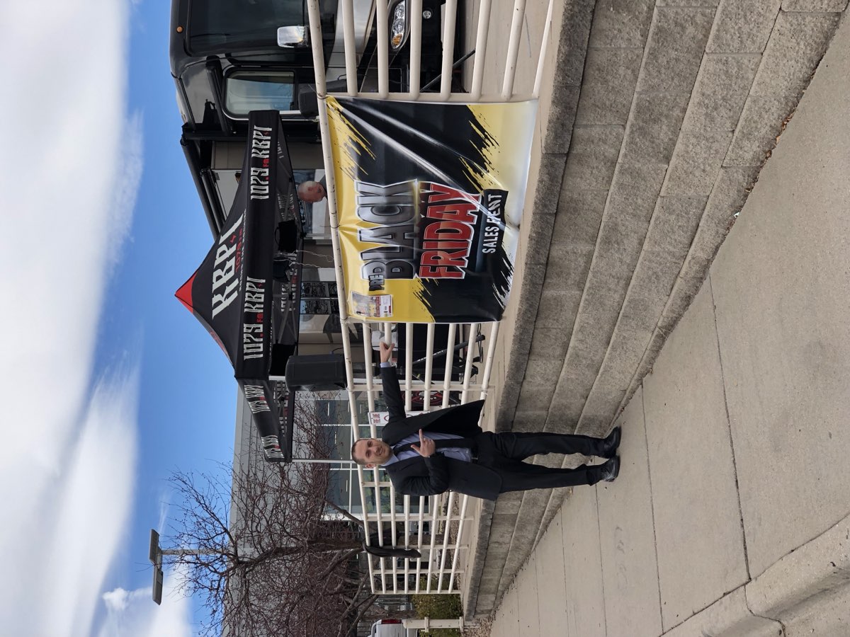 Steve Temes standing beside a Black Friday sales event banner with a radio station's remote broadcast bus parked behind him.